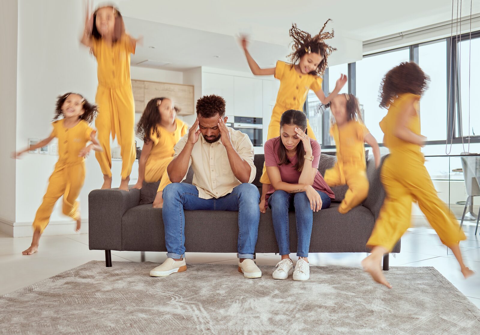Two tired parents on the sofa while their child bounces around the living room, full of energy